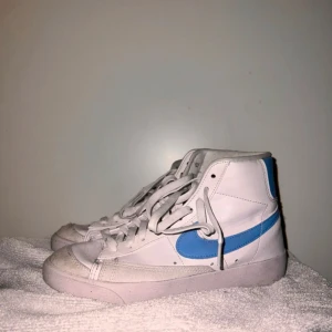 Nike mid Blazer 77 - Säljer ett par klassiska Nike Blazer sneakers i vitt med blå detaljer. Skorna har en retro look med hög snörning och är perfekta för både vardag och gym. De är i bra skick och redo för nya äventyr. Passar perfekt till jeans eller shorts!             Anvönds några gånger i gymmet men annars är dom i ett jättebra skick och ett jätte bra pris för dom🤩