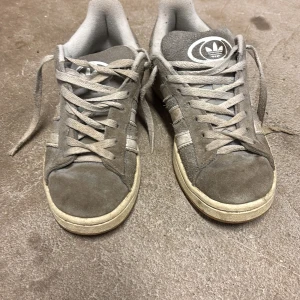 Grå Adidas campus  - Säljer ett par grå Adidas campus i bra skick. De har en klassisk design med vita ränder och snörning. Skorna är gjorda i mocka och har en gummisula som ger bra grepp. Perfekta för vardagsbruk och passar till de flesta outfits.