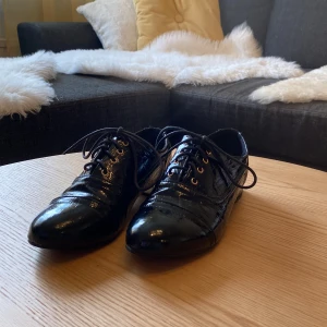 Svarta lackskor med snörning - Snygga svarta lackskor från Yes Mile i mycket bra skick. De har en klassisk design med snörning och en elegant, glansig yta. Perfekta för både fest och formella tillfällen. Skorna har en låg klack och en spetsig tå som ger en stilren look.