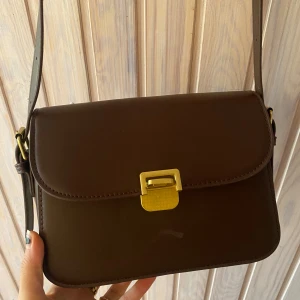 Brun axelväska med gulddetaljer - Säljer en stilren brun axelväska i skinn med justerbar rem och guldfärgat spänne. Väskan har ett huvudfack med dragkedja och en praktisk innerficka. Perfekt för både vardag och fest!