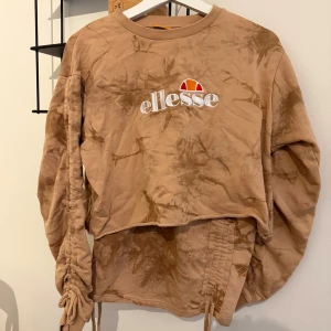 Beige Set med croppad tröja och kjol från Ellesse - Säljer ett set från Ellesse med batikmönster. Den har snörning på båda sidor. Tröjan är strl 12 och kjolen 8. Fint skick, knappt använt 