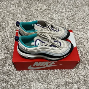 Nike Air Max 97  - Säljer ett par Nike Air Max 97 i vitt med blå och svarta detaljer. Skorna har en ikonisk vågig design och synlig Air-enhet i sulan för extra komfort. De är i bra skick och perfekta för både vardag och träning. Storlek är inte specificerad, men de ser ut att passa en normal passform.