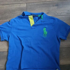 Blå pikétröja Ralph Lauren - Säljer en snygg blå pikétröja från Ralph Lauren i storlek L. Tröjan har en grön broderad logga på bröstet och en orange siffra '3' på ärmen. Den har en gul knappslå med blå knappar.