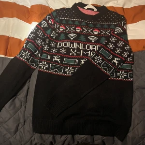Svart jultröja med mönster - Säljer en svart jultröja med färgglatt mönster i rött, grönt och vitt. Tröjan har långa ärmar och ett festligt mönster med snöflingor och andra julmotiv. Perfekt för julfirandet eller en mysig vinterdag!