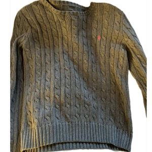 Grå stickad  tröja från Ralph Lauren - Säljer en snygg grå kabelstickad tröja från Ralph Lauren. Den har en klassisk design med rund hals och det ikoniska röda logotypbroderiet på bröstet. Perfekt för kyliga dagar och passar både till vardags och lite finare tillfällen. Supermysig och stilren!