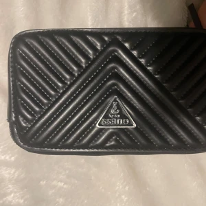 Svart quiltad axelväska från Guess - Snygg svart axelväska från Guess i quiltat skinn. Väskan har en justerbar axelrem med Guess-logga och en praktisk dragkedja. Perfekt för både vardag och fest!