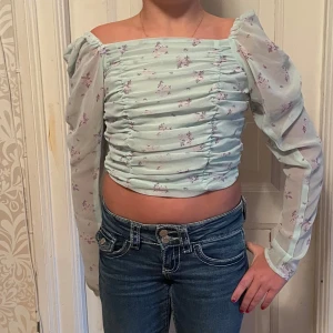 Blommig offshoulder topp - Söt offshoulder topp i ljusgrön med blommigt mönster. Toppen har långa, puffiga ärmar i ett transparent material och en rynkad design framtill. Perfekt för vår och sommar!