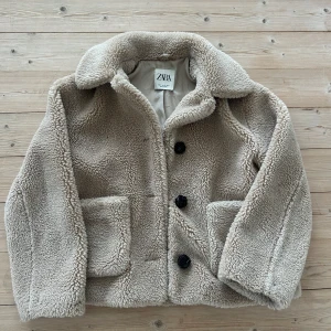 Beige teddyjacka från Zara - Supermysig beige teddyjacka från Zara i storlek XS.Perfekt för höst och vinter med sina långa ärmar och stora knappar. Jackan har en klassisk krage och två praktiska fickor framtill. Den är i mycket bra skick och redo att hålla dig varm och stilren i kylan!
