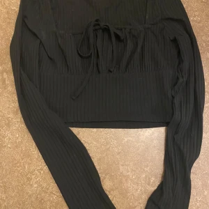 Svart ribbad tröja - Säljer en svart ribbad tröja med långa ärmar och knytning framtill. Perfekt för höst och vinter, den är både stilren och bekväm. Passar bra till både vardag och fest. Tröjan är i bra skick och redo för en ny ägare!