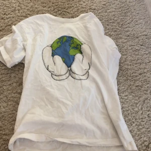 Vit t-shirt med jordmotiv från Uniqlo - Säljer en vit t-shirt från Uniqlo i samarbete med KAWS. T-shirten har ett coolt tryck på ryggen med en jordglob som hålls av två händer. På insidan av kragen står det 'PEACE FOR ALL' och 'KAWS'. Perfekt för en avslappnad och stilren look. Den är i bra skick och passar perfekt till vardags.