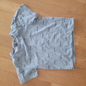 Grå t-shirt från Adidas - Säljer en grå t-shirt från Adidas med deras logga tryckt över hela tröjan. Den är kortärmad och i bra skick. Perfekt för vardagsbruk eller träning. Passar både killar och tjejer. Skicka ett meddelande om du är intresserad! 😊
