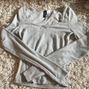 Grå ribbad tröja från H&M Divided - Säljer en superfin grå ribbad tröja från H&M Divided. Den är i storlek XS och har långa ärmar. Tröjan har en söt rosett framtill och spetskant. Jag har klippt av sidenbanden som brukar sitta över axeln då den kliade. Inga fläckar.