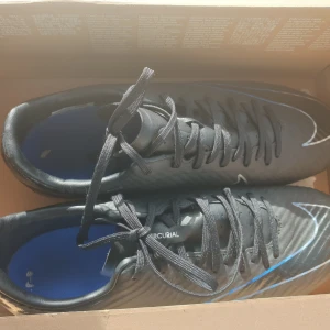 Svarta Nike Mercurial fotbollsskor - Säljer ett par svarta Nike Mercurial fotbollsskor i nyskick. Skorna har en snygg design med blå detaljer och snörning. De är perfekta för fotbollsträning och matcher. Sulan har bra grepp och är designad för att ge optimal prestanda på planen.