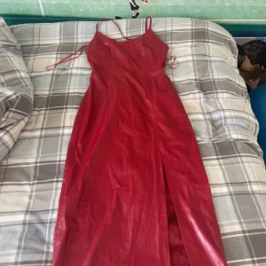 Röd klänning med slits - Snygg röd klänning i syntetmaterial med smala axelband och en elegant slits framtill. Perfekt för festliga tillfällen! Klänningen har en dragkedja bak för enkel påklädning.