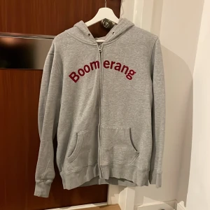 Grå hoodie från Boomerang - Säljer en grå hoodie från Boomerang i storlek L. Den har en dragkedja framtill och en mysig huva. Perfekt för en avslappnad stil och passar bra till både höst och vår. Boomerang-loggan är tryckt i rött på bröstet. Skön och stilren design!