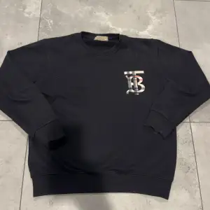 Säljer en snygg svart sweatshirt från Burberry i storlek S. Tröjan har ett coolt mönstrat TB-tryck på bröstet och en röd broderad detalj på ärmen. Perfekt för både höst och vår, och den är superbekväm att ha på sig. Passar perfekt till jeans eller chinos för en stilren look.