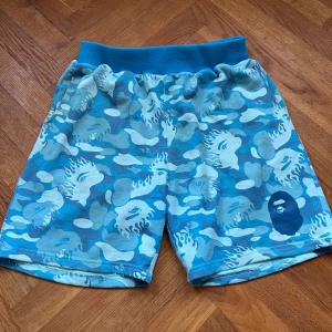 BAPE Camo Shorts - Säljer ett par blå camo shorts från A Bathing Ape i storlek M. De har ett coolt kamouflagemönster i olika blå nyanser och en elastisk midja för bekväm passform. Fick dem i present för 3 år sen så inte helt säker på autenciteten. Perfekta för sommarens alla äventyr! De har lite fläckar men fortfarande bra skick och inga hål, lite noppriga.