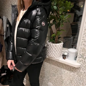 Svart pufferjacka från Colmar - Säljer en snygg svart pufferjacka från Colmar. Den är perfekt för kalla dagar med sin tjocka vaddering och har en praktisk huva. Jackan har en glansig finish och stängs med dragkedja. Perfekt för vintern! ❄️