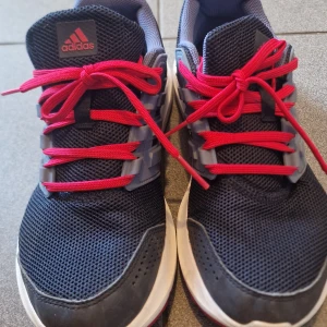 Adidas sneakers med röda snören - Säljer ett par Adidas sneakers i mycket bra skick. De är mörkblå med röda snören och har en vit sula. Perfekta för träning eller vardagsbruk. Skorna är bekväma och har en snygg design med Adidas-loggan på plösen.