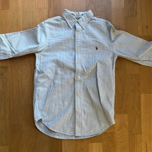 Randig skjorta från Ralph Lauren - Säljer en snygg och klassisk randig skjorta från Ralph Lauren i blått och vitt. Skjortan har långa ärmar och knappar framtill. Perfekt för både vardag och fest! 🕺
