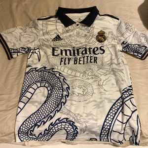 Real madrid X Dragon T-shirt - Säljer en snygg vit Real Madrid tröja från Adidas med ett coolt drakmönster i grått. På baksidan av tröjan står det ”Vini jr” med nr 20.Tröjan har korta ärmar och klubbens emblem på bröstet. Den är i mycket bra skick och perfekt för alla fans! 🐉⚽nypris:500kr. Använd ca 3-5 gånger