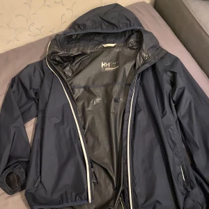 Mörkblå regnjacka från Helly Hansen - Säljer en mörkblå regnjacka från Helly Hansen i bra skick. Jackan har en praktisk huva och dragkedja framtill. Perfekt för regniga dagar och håller dig torr och bekväm. Passar både till vardags och utomhusaktiviteter.