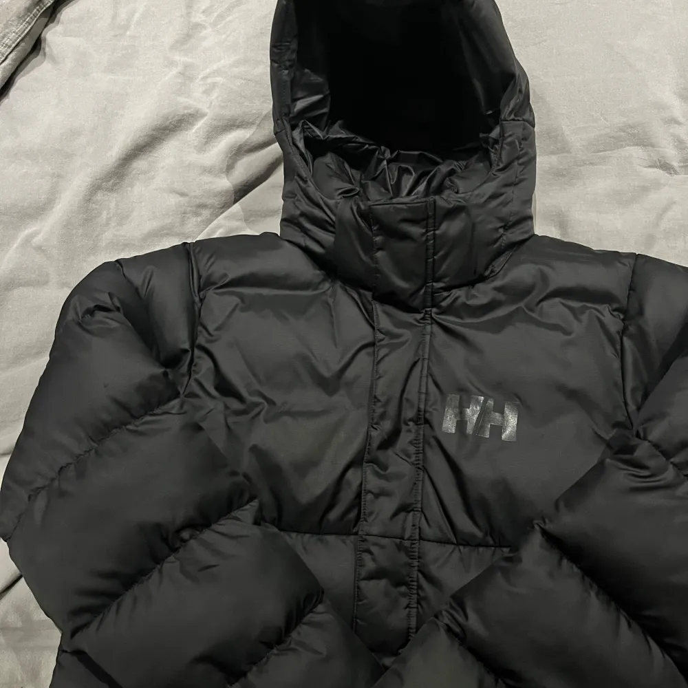 Säljer en svart dunjacka från Helly Hansen i storlek 164. Jackan är perfekt för kalla vinterdagar med sin tjocka vaddering och huva. Den har en stilren design med Helly Hansen-logga på bröstet och ryggen. Jackan är i mycket bra skick och redo för nya äventyr! 🖤Luvan är avtagbar. . Takit.
