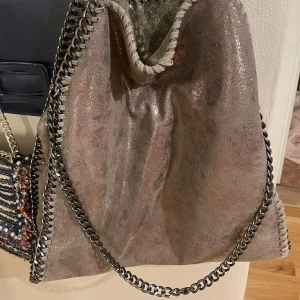 Glittrig axelväska med kedjedetaljer - Snygg glittrig axelväska i en skimrande grå färg. Väskan har coola kedjedetaljer längs kanterna och en kedja som axelrem. Perfekt för att ge lite extra bling till din outfit!