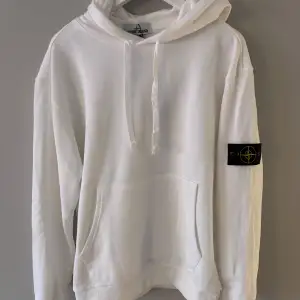 Säljer en snygg vit hoodie från Stone Island. Den har en klassisk design med en stor ficka fram och en justerbar huva. På ärmen finns det ikoniska Stone Island-märket. Perfekt för både vardag och chill.