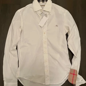 Burrbery skjorta - Säljer en stilren vit skjorta från Burberry i bomull. Den har långa ärmar och klassiska knappar framtill. Detaljerna på insidan av kragen och ärmsluten är i Burberrys ikoniska rutmönster. Perfekt för både vardag och fest!