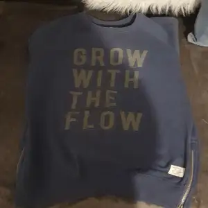 Säljer en blå sweatshirt med texten 'GROW WITH THE FLOW' tryckt på framsidan. Tröjan har dragkedja på båda sidorna och är i bra skick. Perfekt skoltröja