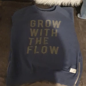 Blå sweatshirt med text - Säljer en blå sweatshirt med texten 'GROW WITH THE FLOW' tryckt på framsidan. Tröjan har dragkedja på båda sidorna och är i bra skick. Perfekt skoltröja