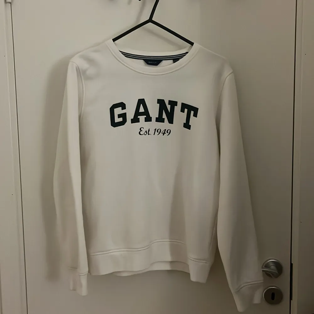 Säljer en stilren vit sweatshirt från GANT med mörkgrön text 'GANT Est. 1949' på framsidan. Tröjan har långa ärmar och en rund halsringning. Perfekt för en avslappnad stil.. Neuletakit & Villapaidat.