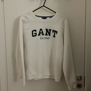Vit sweatshirt från GANT stl.164/xs - Säljer en stilren vit sweatshirt från GANT med mörkgrön text 'GANT Est. 1949' på framsidan. Tröjan har långa ärmar och en rund halsringning. Perfekt för en avslappnad stil.
