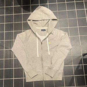 Grå hoodie från Polo Ralph Lauren - Säljer en stilren grå hoodie från Polo Ralph Lauren med dragkedja och vit dragsko. Den har en liten grön logga på bröstet och är tillverkad i mjukt material. Perfekt för en avslappnad look.