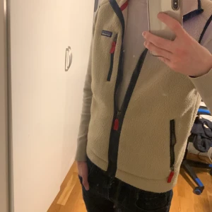 Beige fleeceväst från Patagonia - Säljer en beige fleeceväst från Patagonia med dragkedja och röda detaljer. Västen har en bröstficka och två sidofickor med dragkedjor. Perfekt för lager-på-lager under kyliga dagar. Köpt för 1300