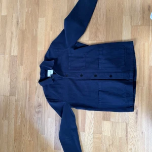 Mörkblå overshirt från A Day's March - Snygg mörkblå overshirt från A Day's March med knappar framtill och två stora fickor. Perfekt för lager-på-lager-stil. Den har en klassisk krage och är tillverkad i ett mjukt material.