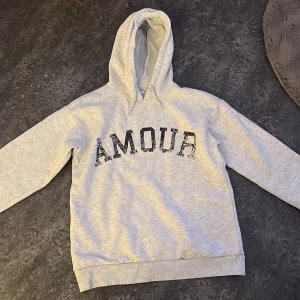 Grå hoodie från Gina Tricot med tryck - Säljer en grå hoodie från Gina Tricot med texten 'AMOUR' på framsidan och en stjärna på baksidan. Tröjan har en klassisk huva och långa ärmar. Perfekt för en avslappnad stil. 🩶 Men liten fläck på baksidan men går säkert bort