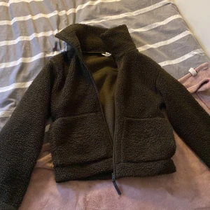 Svart teddyjacka med dragkedja - Mysig svart teddyjacka med dragkedja framtill och hög krage. Perfekt för kyliga dagar när du vill hålla dig varm och bekväm. Jackan har långa ärmar och två praktiska fickor framtill.