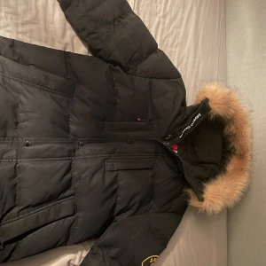 Svart dunjacka med pälsdetalj från Zavetti Canada - Snygg svart dunjacka från Zavetti Canada med pälsdetalj på huvan. Jackan har en dragkedja och knappar framtill samt flera praktiska fickor. Perfekt för kalla vinterdagar.