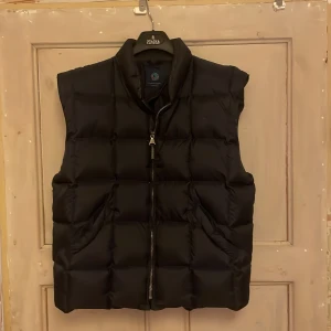 Svart dunväst från Boomerang - Säljer en svart dunväst från Boomerang i storlek medium. Västen har en dragkedja framtill och är tillverkad av 100% nylon med fyllning av 90% ankdun och 10% fjäder. Perfekt för kyligare dagar!