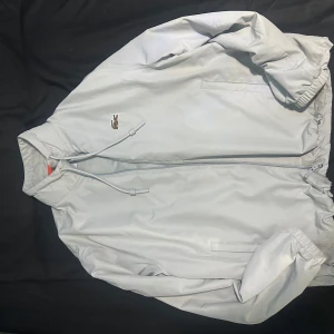 Vit vindjacka från Lacoste - Säljer en stilren vit vindjacka från Lacoste med dragkedja och elastiska muddar. Jackan har en broderad logotyp på bröstet och är perfekt för vår och sommar. Den är tillverkad i ett lätt syntetmaterial och har en normal passform.