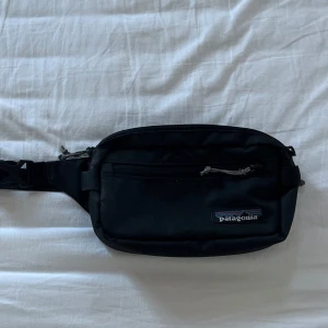 Svart midjeväska från Patagonia - Säljer en praktisk svart midjeväska från Patagonia. Väskan har en justerbar rem och dragkedja för säker förvaring. Den är i bra skick utan skador och defekter