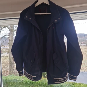 Marinblå jacka från Tommy Hilfiger - Snygg marinblå jacka från Tommy Hilfiger med dragkedja och knappar. Jackan har långa ärmar och märkesdetaljer vid fållen och ärmsluten. Perfekt för en stilren look.