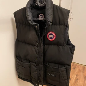Canada Goose väst - Säljer en svart dunväst från Canada Goose med en broderad logotyp på bröstet. Västen har en dragkedja och knappar framtill samt två stora fickor. Perfekt för kyligare dagar.