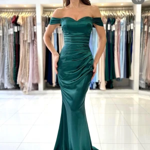 Grön balklänning  - Elegant grön långklänning i satin med offshoulder-design. Klänningen har en figurnära passform och en vacker drapering framtill. Perfekt för speciella tillfällen. Använd 1 gång på balen. Lite sliten längs ner samt en fläck som knappt syns. Är 163, hade klackar som var 11 cm men klänningen var fortfarande lite för lång. Går att sy på. Från dressroom linne. Nypris 4000kr