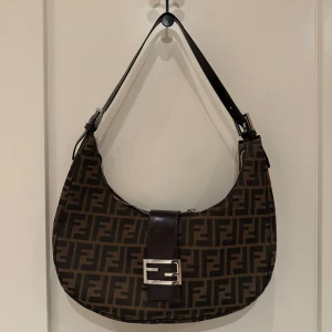 Brun axelväska  - Snygg brun axelväska med monogrammönster och en stilren metallspänne framtill. Väskan har en justerbar axelrem och dragkedja upptill. Köpt här på Vinted, dustbag medföljer. Har inte kvitto för garanti/äkthet