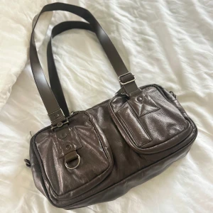 Brun mandarina duck axelväska  - Snygg brun axelväska i skinn med två framfickor och justerbara axelremmar. Väskan har detaljer som spännen och en D-ring framtill. Perfekt för att bära med sig det nödvändigaste med stil.