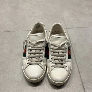 Vita Gucci ace skor - Säljer ett par vita sneakers från Gucci med klassiska röd och grön rand på sidan och en broderad bi-detalj. Skorna har snörning och är tillverkade i skinn. Baktill finns en röd och en grön hälflik.