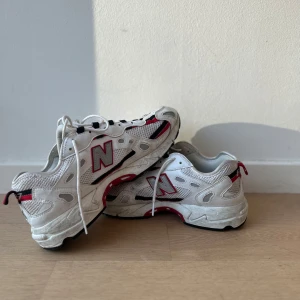 Vita sneakers från New Balance - Säljer ett par vita sneakers från New Balance med röda och svarta detaljer. Skorna har snörning och en bekväm sula, perfekt för vardagsbruk. De har en sportig design med märkets logotyp på sidan.